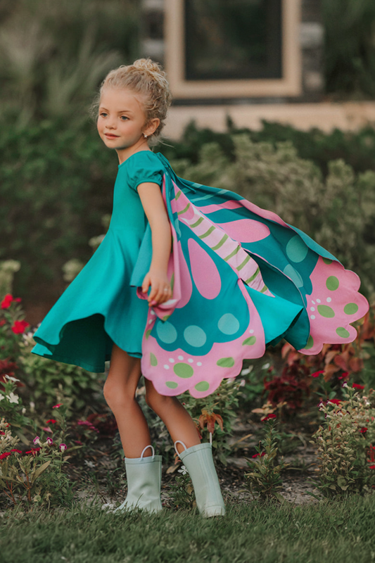 A young girl twirls in a beautiful turquoise dress. The short puff sleeves and flowing skirt look classic. There is a beautiful butterfly wing cape that attaches with buttons in the back. The butterfly has bright teal, pink and green designs on the wings.