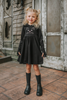 A young girl is wearing a black long sleeve cotton twirl dress. There is a kitty embroidered on the chest. The sleeves have pink kitty paws on them with thumbholes to put your thumb through. Perfect for spooky season.