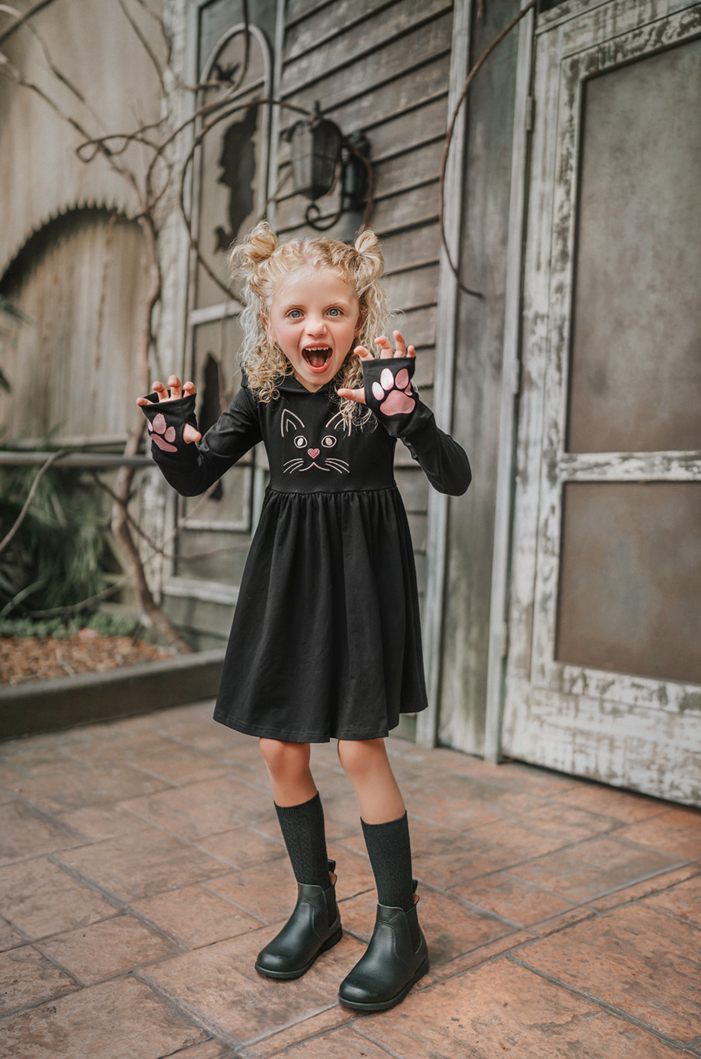 A young girl is wearing a black long sleeve cotton twirl dress. There is a kitty embroidered on the chest. The sleeves have pink kitty paws on them with thumbholes to put your thumb through. Perfect for spooky season.