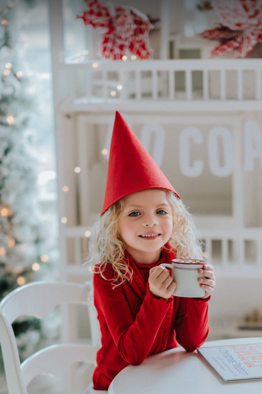 A young child wearing the Red Elf Hat, a red outfit with a white collar, and holding holiday lights, smiles while looking up. The hat is a cone shape and true red in color. The background is blurred, emphasizing the child's joyful expression and festive fun, radiating Christmas cheer.