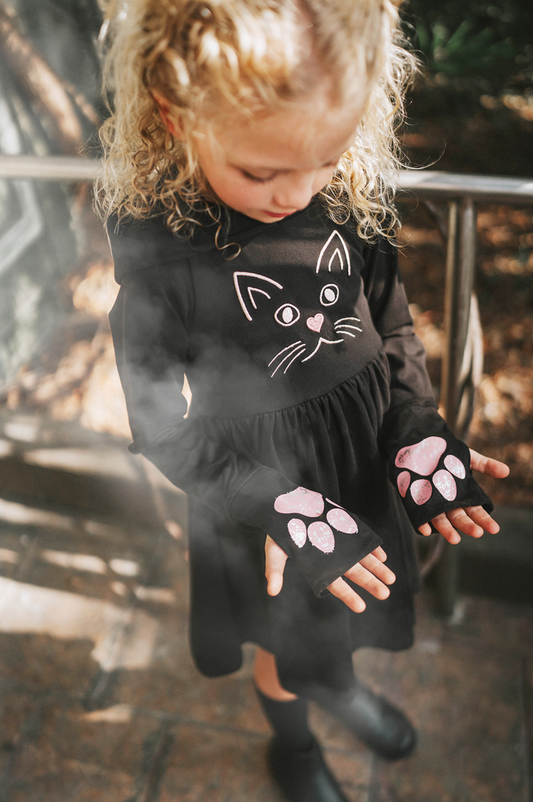 A young girl is wearing a black long sleeve cotton twirl dress. There is a kitty embroidered on the chest. The sleeves have pink kitty paws on them with thumbholes to put your thumb through. Perfect for spooky season.