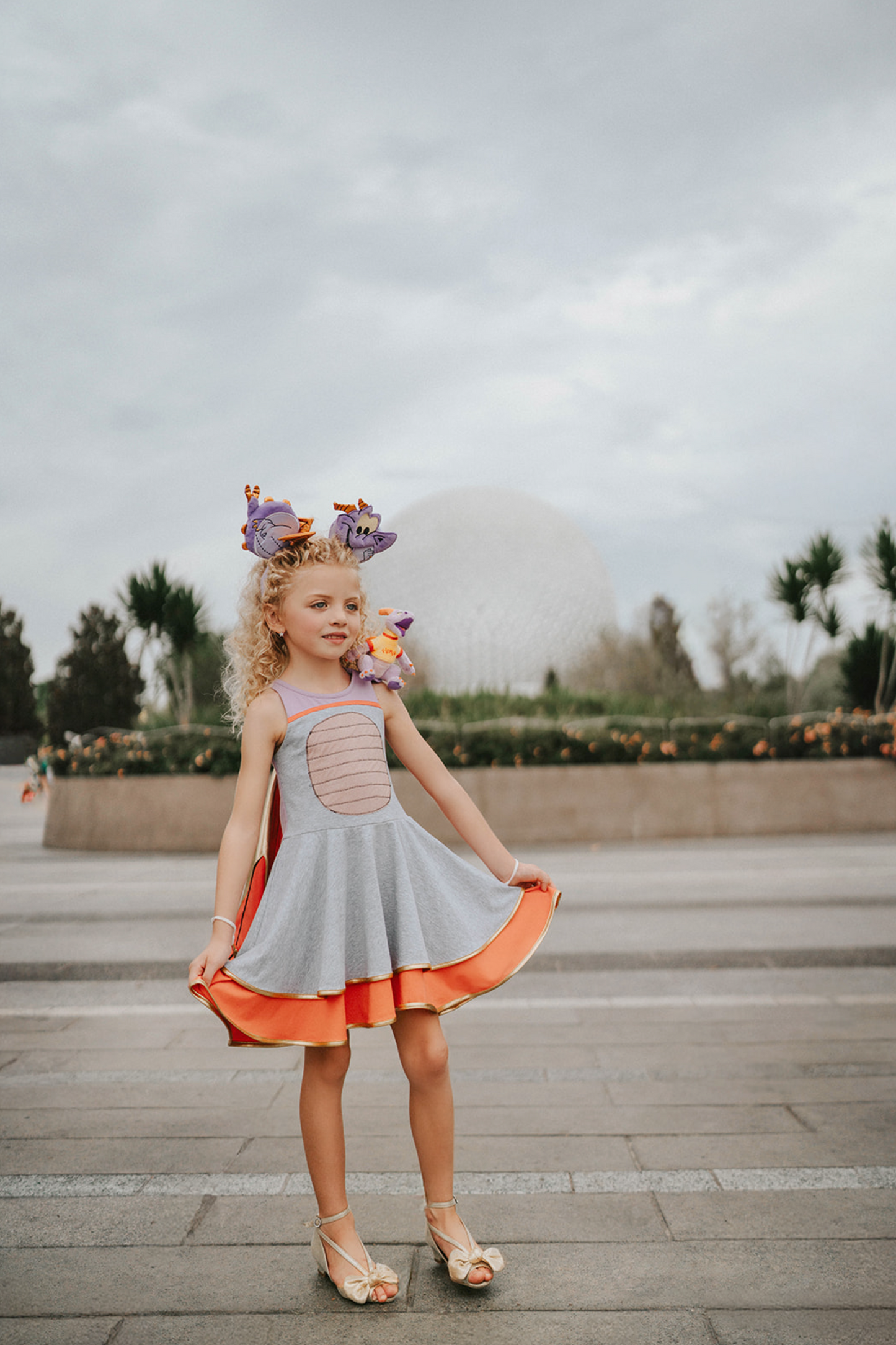 A young girl is wearing a light purple and heather grey cotton dress. There is a light pink sparkle oval to resemble a belly. The skirt is a double twirl with the second layer in orange. The detachable wings are gold and orange.