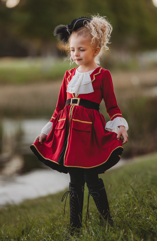 A young girl stands outdoors, wearing a vibrant red Pirate Twirl Dress with gold trim with a white ruffled neckline. Adorned with delicate ruffle detailing at the sleeves and a black waistband with gold buckle.