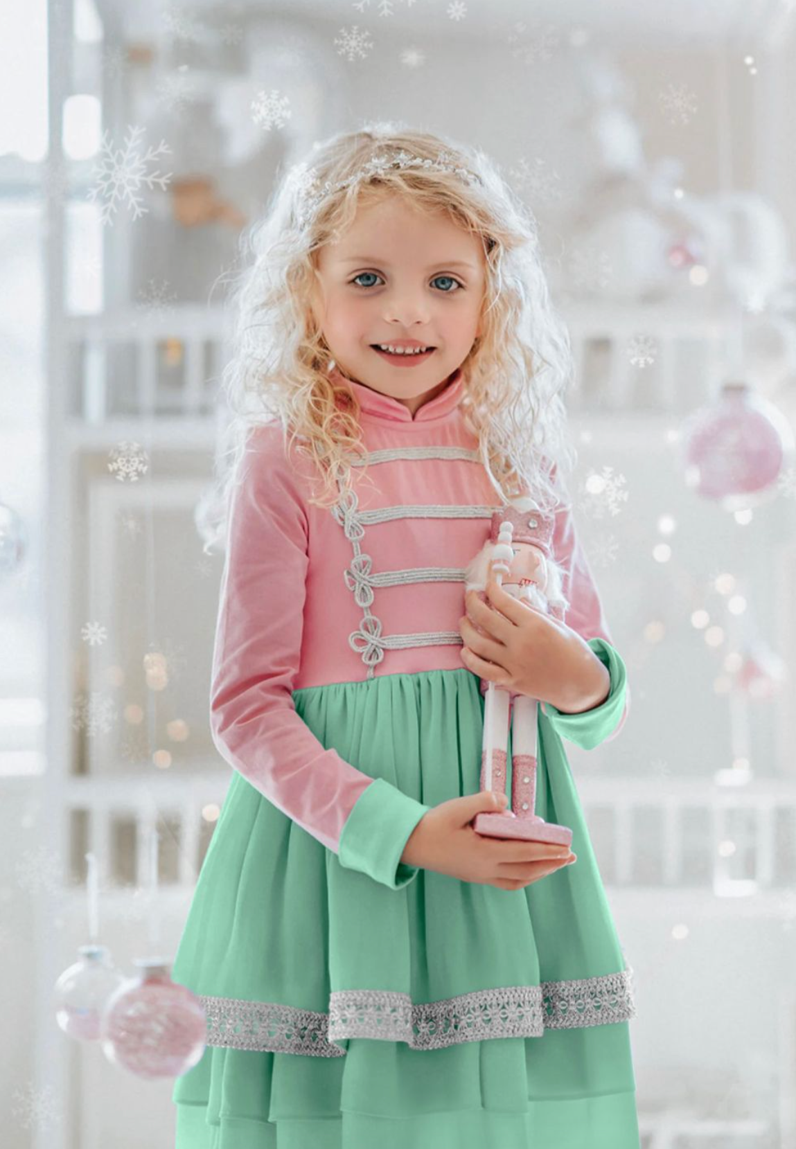 A girl in a pastel pink and mint green twirl dress with silver braiding, holding a toy soldier, with a blurred Christmas-themed backdrop.