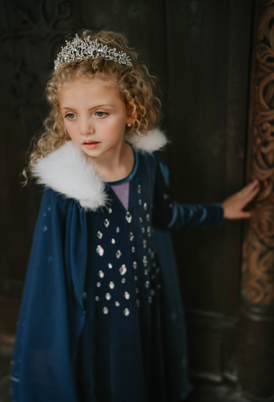 A young girl is wearing a navy blue chiffon cape with white faux fur trim. The matching velvet gown is adorned with diamond shape crystal gems of various sizes. There is a small lavender triangle near the neck.