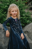 A young girl is wearing a navy blue velvet gown is adorned with diamond shape crystal gems of various sizes. There is a small lavender triangle near the neck.