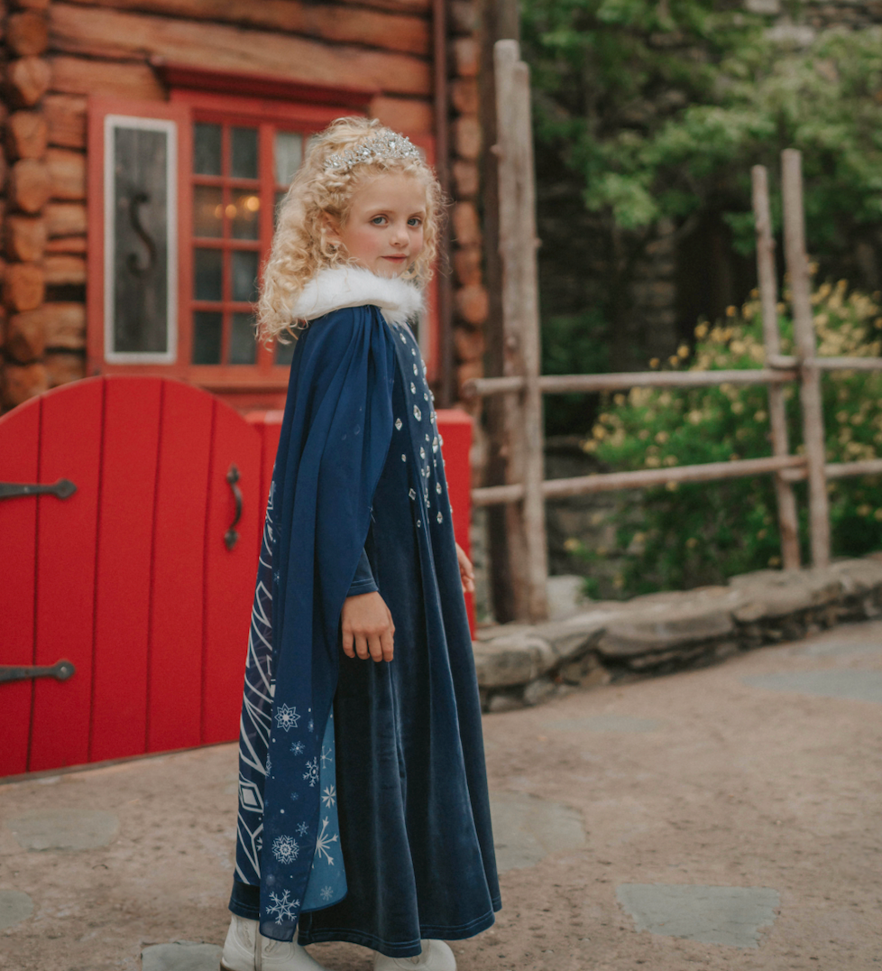 A young girl is wearing a navy blue velvet gown is adorned with diamond shape crystal gems of various sizes. There is a small lavender triangle near the neck. She has on a navy blue chiffon cape with white faux fur trim on the collar. There is a snow flake design on the back of the cape.