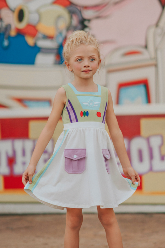 A young girl wears a sporty space hero twirl dress. The cotton dress is sleeveless and has light green panels on the bodice with embroidered button like features. There are light green and light purple racing stripes down the side. The front has two pockets on the skirt.