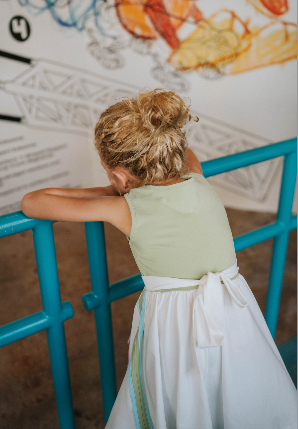 A young girl wears a sporty space hero twirl dress. The cotton dress is sleeveless and has light green panels on the bodice with embroidered button like features. There are light green and light purple racing stripes down the side. The front has two pockets on the skirt.
