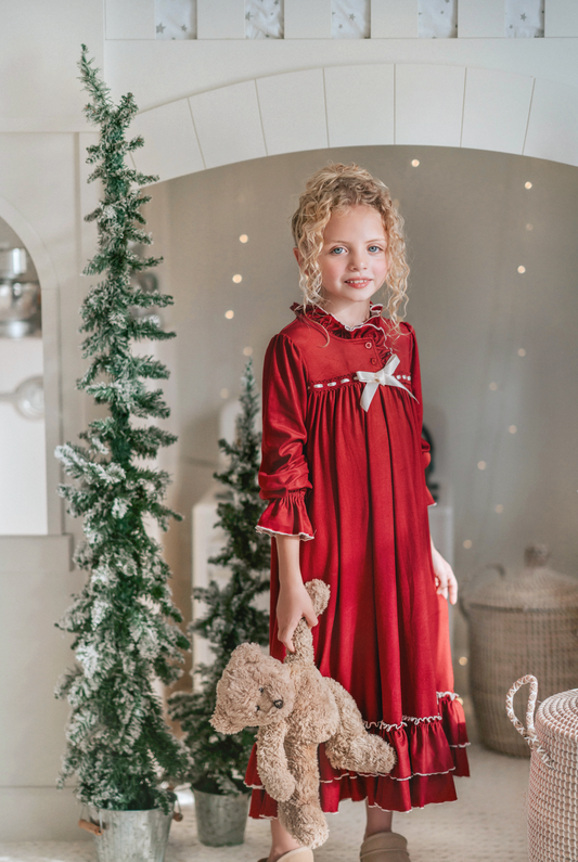 A young girl is wearing The Original Clara Nutcracker Gown in Merry Maroon, featuring an ivory bow. The buttery soft fabric is cozy and sensory friendly. The long sleeves and the ruffles at the hem add a traditional classic look.