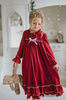 A young girl is wearing The Original Clara Nutcracker Gown in Merry Maroon, featuring an ivory bow. The buttery soft fabric is cozy and sensory friendly. The long sleeves and the ruffles at the hem add a traditional classic look.