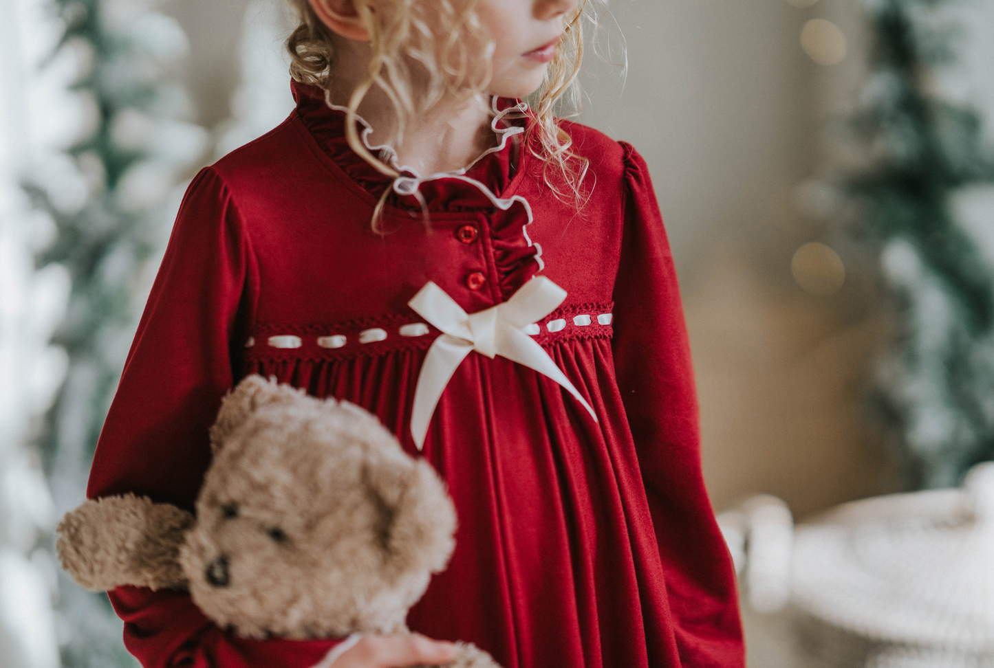 A young girl is wearing The Original Clara Nutcracker Gown in Merry Maroon, featuring an ivory bow. The buttery soft fabric is cozy and sensory friendly. The long sleeves and the ruffles at the hem add a traditional classic look.