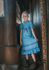 A girl stands in our buttery soft Ice Queen Gown. A blue nightgown with Scandinavian floral print. Short puff sleeves and a ruffle at the hem.