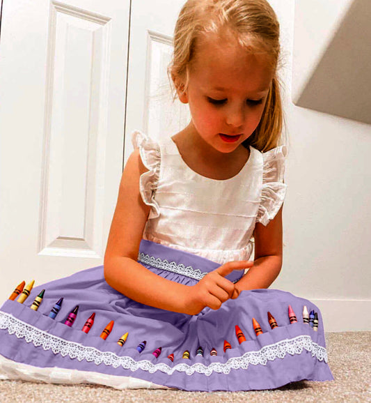 A young child sitting on the floor wearing a white dress and a lavender apron with crochet lace details. The apron has pockets with crayons placed in it.