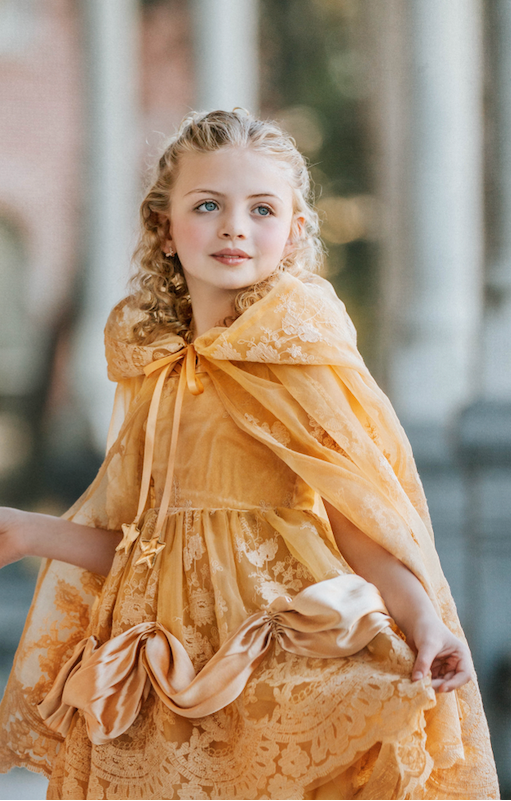 Young girl in a yellow dress with a hood holding a red rose, standing on a street.