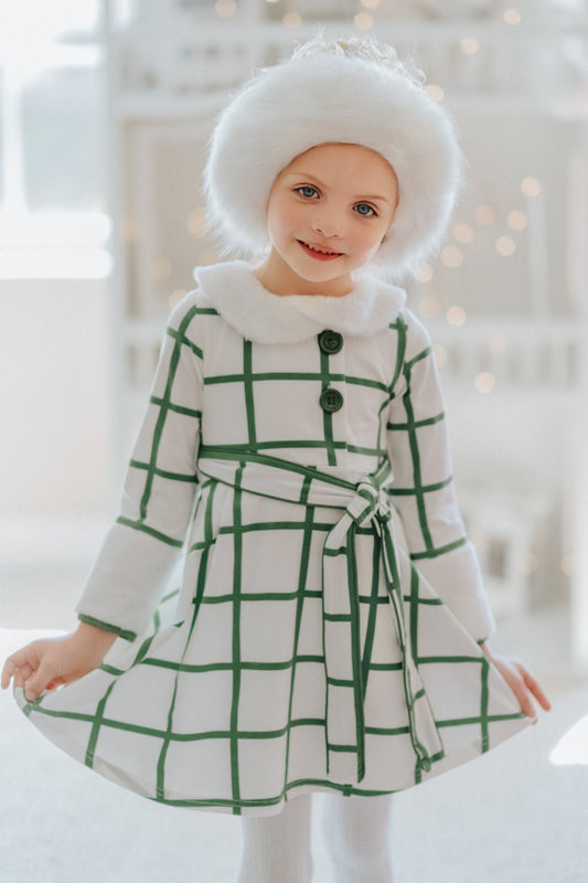A young girl wears a white and green plaid windowpane dress. The dress has green buttons at the top and a belt tie. The collar and cuffs are a soft white fur.