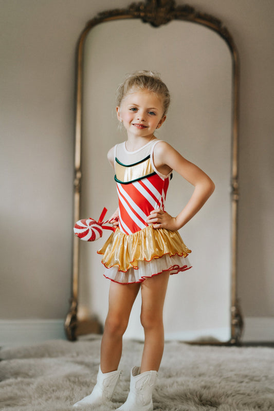 A young girl is wearing a skirted leotard. Red and white candy cane graphic fabric is paired with metallic gold and green velvet accents. The skirt has a metallic gold layer and a white layer below trimmed with red.