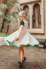 A young girl is wearing our Mummy Twirl Dress. The cotton twirl dress an off white with black short sleeves. There is a black band across the chest with vibrant green eyes. The black lines over the dress create a mummy bandage and there is a green hem. This dress is perfect for spooky season!