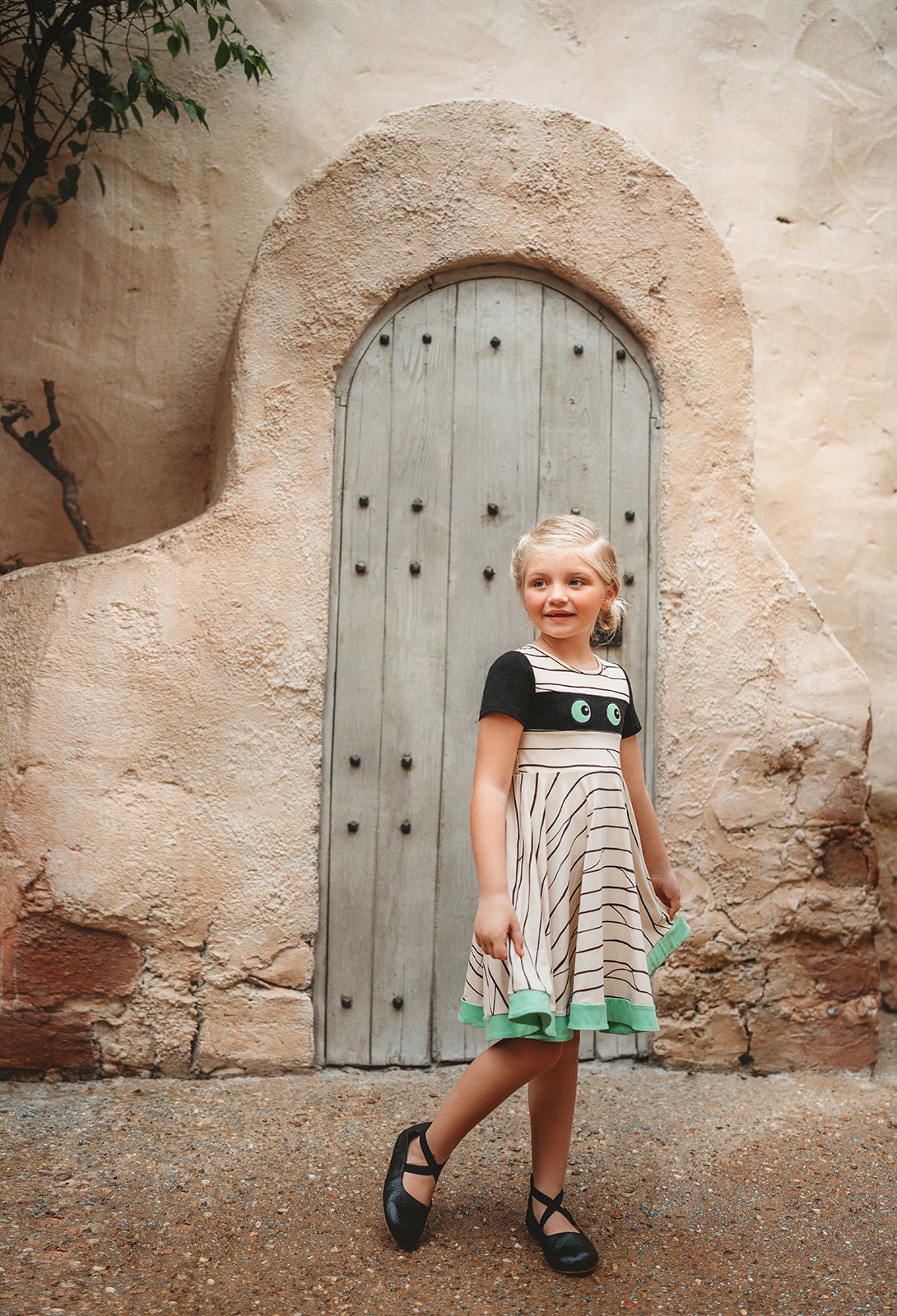 A young girl is wearing our Mummy Twirl Dress. The cotton twirl dress an off white with black short sleeves. There is a black band across the chest with vibrant green eyes. The black lines over the dress create a mummy bandage and there is a green hem. This dress is perfect for spooky season!