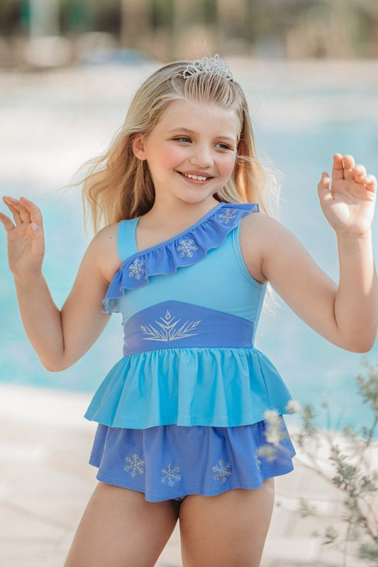 A young girl stands wearing a light blue 2 piece swim set. The top has an asymmetric cornflower blue ruffle with silver snow flake accents. The skirted bottom is cornflower blue and has silver snowflake embroidered around it.