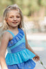 A young girl stands wearing a light blue 2 piece swim set. The top has an asymmetric cornflower blue ruffle with silver snow flake accents. The skirted bottom is cornflower blue and has silver snowflake embroidered around it.