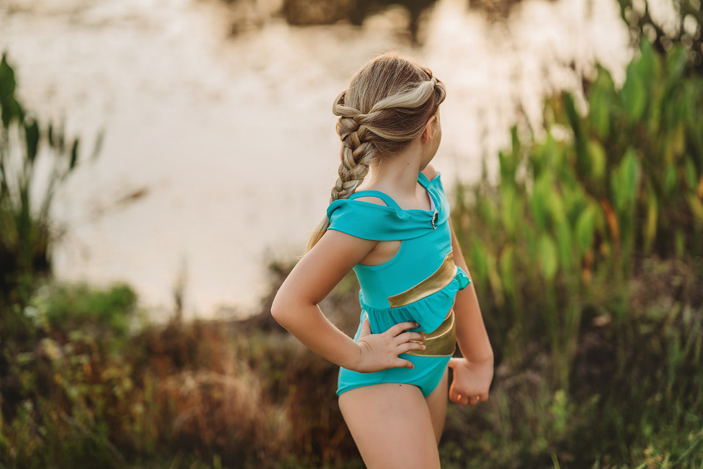 A young girl is wearing a bight teal and gold 2pc swim set. The top has open shoulders and straps. At the hem is a gold band and teal ruffles. There is a teal blue and gold jewel at the neckline. The bottom is a modest cut with gold waist and teal.