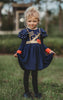 A young girl is wearing a long sleeve navy blue twirl dress. The sleeves have two large flutter ruffles at the shoulder with metal gold dots. The chest is adorned with gold detail and crowns. A red heart with a gold crown in the center is on the lower left chest.