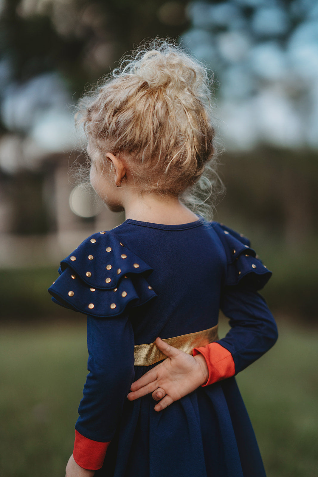 A young girl is wearing a long sleeve navy blue twirl dress. The sleeves have two large flutter ruffles at the shoulder with metal gold dots. The chest is adorned with gold detail and crowns. A red heart with a gold crown in the center is on the lower left chest.