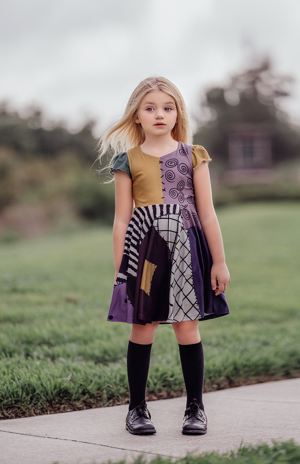 A young girl with long blonde hair is standing on a grassy field, wearing "The Pumpkin King's Friend" dress, its colorful patchwork in purple, black, and green made of soft poly-spandex blend fabric. She pairs the dress with black knee-high socks and looks off to the side while holding the edges of her dress. Trees stand in the background.