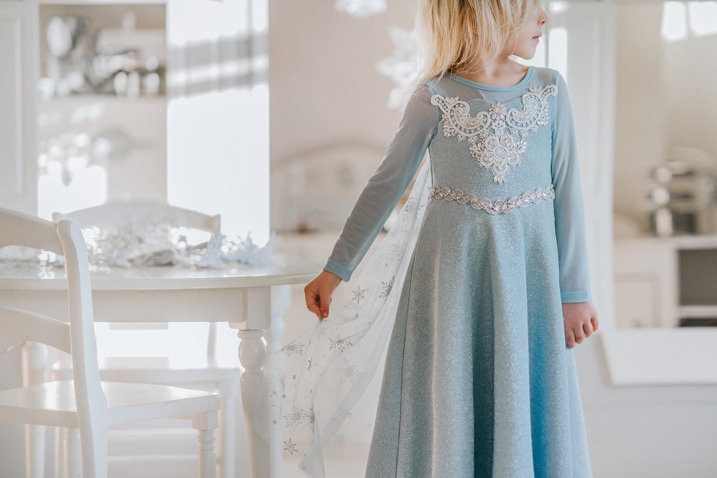 A young girl is dressed in the sparkly light blue Portrait Collection Ice Queen Gown adorned with intricate custom lace at the neckline with rhinestones. A light blue cape with silver sparkle snowflakes is buttoned to the back of the gown.