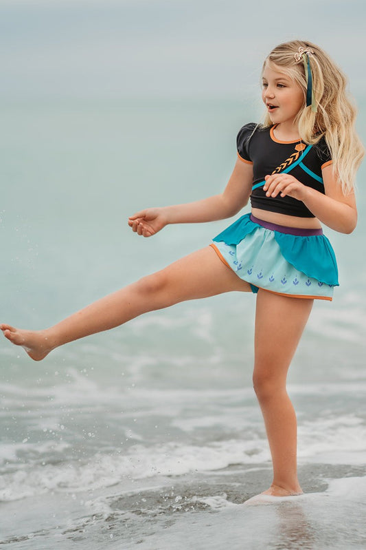 A young girl is wearing a 2 piece swim set. The cropped top is black with turquoise and goldenrod accents and puff sleeves. The bottom has a turquoise ruffle over a mint teal skirt with goldenrod hem accent.
