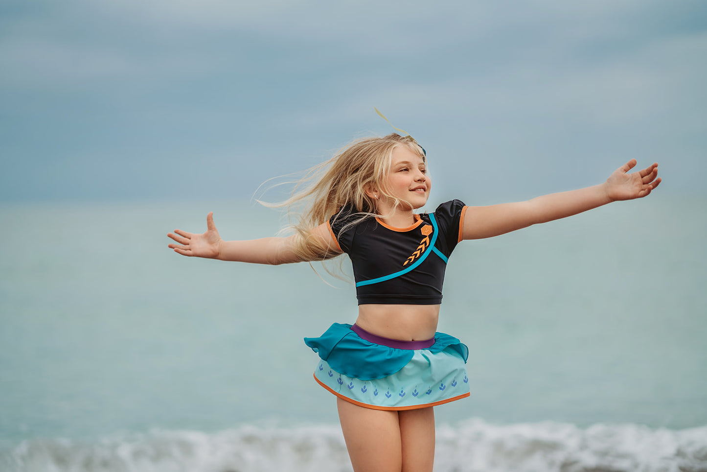 A young girl is wearing a 2 piece long sleeve rash guard swim set. The top is black with turquoise and goldenrod accents. The bottom has a turquoise ruffle over a mint teal skirt with goldenrod hem accent.