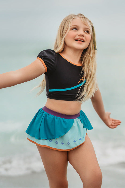 A young girl is wearing a 2 piece long sleeve rash guard swim set. The top is black with turquoise and goldenrod accents. The bottom has a turquoise ruffle over a mint teal skirt with goldenrod hem accent.