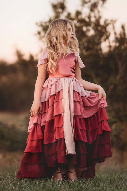 A young girl is wearing the Yule Love all of the ruffles Maxi Dress in shades of pink and red. The soft pink flutter sleeves flow over her shoulder. The bodice is a pink mauve and leads into cascading layers chiffon that start with dusty pink and flow down into a deep maroon.