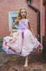 A young girl is adorned in the Portrait Collection Rapunzel Gown—a flowing, light violet princess dress featuring a purple ribbon corset design on the bodice. The skirts are 3 layers of light dusty purple, pink and lavender. The skirt is perfect for twirling and dancing in any enchanted land.