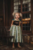 A young girl is wearing a Portrait Collection Coronation Ice Princess Gown inspired by Princess Anna from the movie "Frozen." The dress showcases a black velvet bodice with custom embroidery and a skirt adorned with green and gold accents. The skirt is shimmering in shades of green with embroidered designs along the panels. There is tulle with cotton lining underneath to add fullness to the skirt.