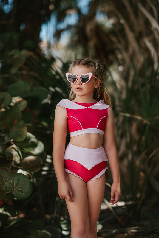 A young girl is wearing a pink 2 piece swimsuit. The Sleeping Beauty swim set has a pink collar that resembles a ballgown. The top has light pink piping and trim. The modest bottoms have a pink overlay and ruffles on the legs.