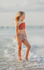 A young girl stands at the edge of the sea, smiling. She's wearing a bright orange and red patterned top and a grey swimsuit bottom adorned with a white hibiscus flower—it's the Polynesian Princess Two Piece Swimsuit made from SPF 50 material. The gentle waves touch her feet, blending with sky hues in the background.
