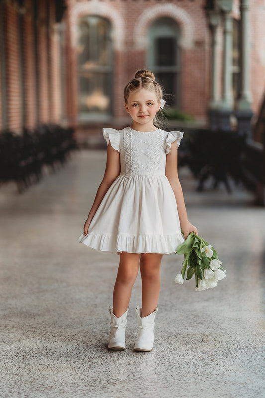 A young girl is wearing the Ethel Fancy Pinafore in Soft White with ruffled sleeves. There is a matching lace overlay on the bodice. A pettiskirt can be added underneath the dress for fullness.