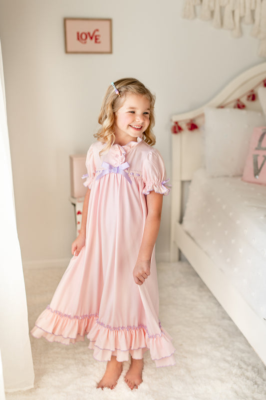 A young girl is wearing The Original Clara Nutcracker Gown in Blush, featuring blush pink ruffles and a lavender bow. The buttery soft fabric is cozy and sensory friendly. The puff sleeves with ribbon detail and the ruffles at the hem add a traditional classic look.