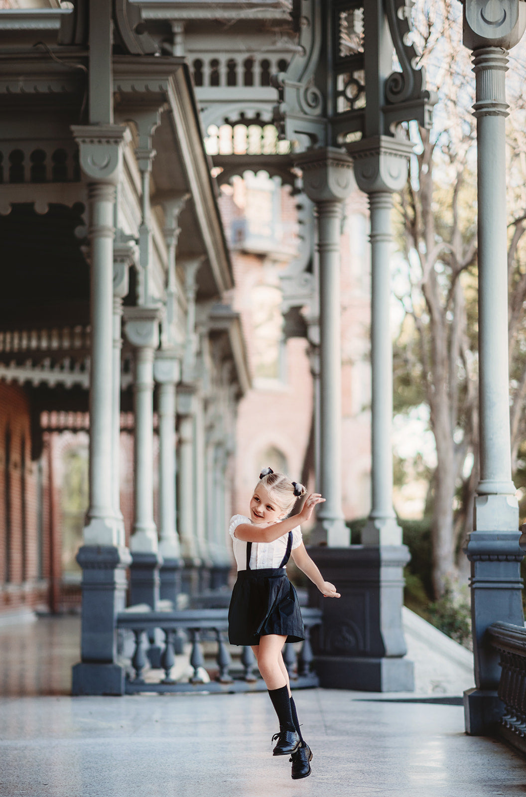 A young girl wears the Plaza Girl Everyday Dress—a white button-down shirt with ruffle accents and short sleeves tucked into a knee-length black skirt with suspenders made of high-quality woven cotton.