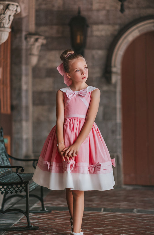 A young girl is wearing The Fairy Godmother Collection Cinderelly Pink Dress. The dress is woven cotton with a white collar and pink bow. The full skirt has bows at the bottom with a white hem.