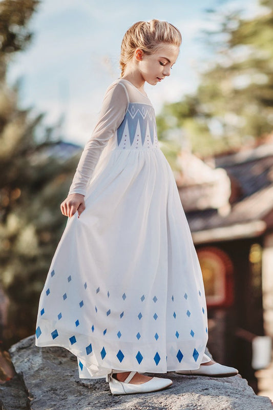 A young girl is wearing the Queen of the Forest Maxi Dress adorned with blue diamond patterns along the bottom. The maxi dress also features a geometric design on the upper bodice and white mesh sleeves and neckline. The removable cape is fastened to the back with buttons.