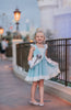A young girl in an "Our Original Cinderella Twirl Dress" stands near a castle. The dress has a white flutter ruffle on the straps. There is a white skirt ruffle that overlays the blue twirl skirt. The soft cotton material is comfortable and breathable.