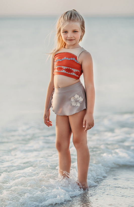 A young girl stands at the edge of the sea, smiling. She's wearing a bright red swim top with flower and pattern band. The tan-grey swimsuit bottom is adorned with a tan hibiscus flower—it's the Polynesian Princess Two Piece Swimsuit made from SPF 50 material. The gentle waves touch her feet, blending with sky hues in the background.