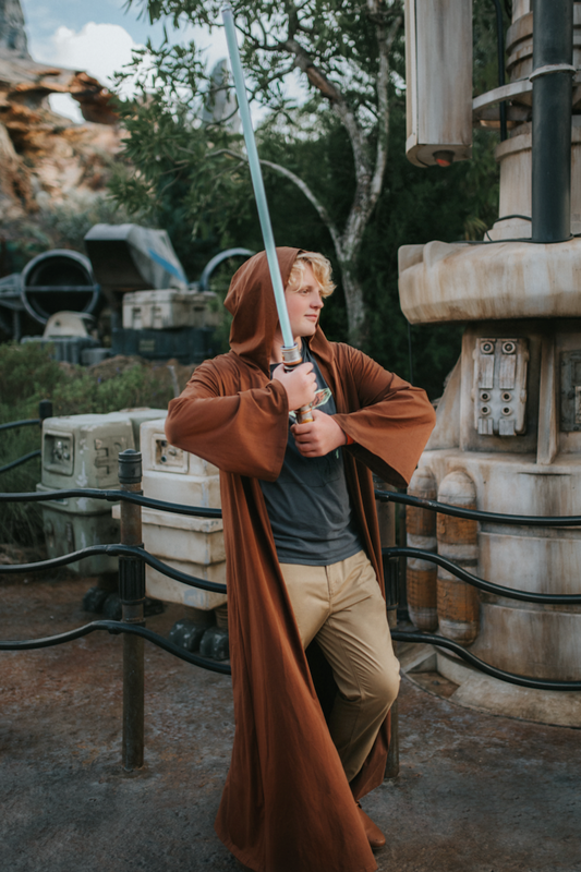 A young teen boy is wearing a sienna brown robe with hood wielding a light saber. The robe is open and has large sleeves. The robe ankle-floor length.