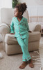 A young child is wearing the Gingham Plaid Unisex two-piece pajama set, featuring a green and white checkered pattern with red piping and white buttons. Ideal for Christmas family photos.