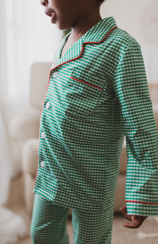 A young child is wearing the Gingham Plaid Unisex two-piece pajama set, featuring a green and white checkered pattern with red piping and white buttons. Ideal for Christmas family photos.