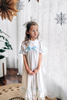 A young girl is wearing The Original Clara Nutcracker Gown in White, featuring white ruffles and a light blue bow. The buttery soft fabric is cozy and sensory friendly. The puff sleeves with ribbon detail and the ruffles at the hem add a traditional classic look.