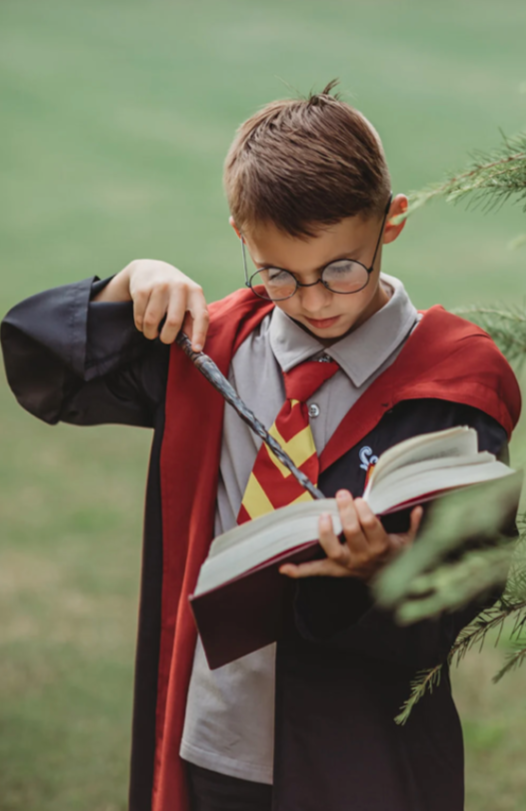 A young boy dressed in the Wizard Ensemble, featuring a red and yellow striped tie, and a black cotton jersey knit robe reminiscent of the Harry Potter series. The robe is lined with brick red and there is a logo sewn over the left chest.
